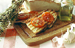 Bruschetta mit Tomaten auf einem Holzbrett, umgeben von Thymian, einem angeschnittenen Laib Brot, Oliven&ouml;l und Knoblauch