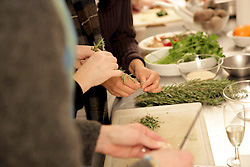 Die H&auml;nde von drei Personen zupfen und schneiden Kr&auml;uter, im Hintergrund Sch&uuml;sseln mit weiteren Zutaten