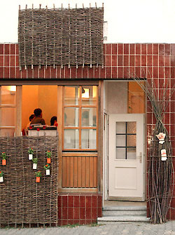 Blick von au&szlig;en auf den Eingang und durch das offene Fenster auf eine Gesellschaft im warm erleuchteten Speiseraum von pane e rose